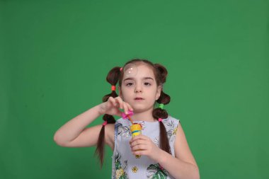 Sekiz yaşında, komik saç stili, çiçekli mavi bir bluz giyen, elinde bir şişeyle oynayan, sabun köpüğü üfleyen, çocukların eğlencesine katılan komik bir kız.