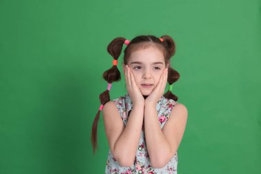 Havalı, genç ve güzel bir kız, sekiz yaşında, harika bir ruh hali içinde, mavi bluzlu, çiçekli, parlak yeşil bir arka planda hoş bir saç stili var.