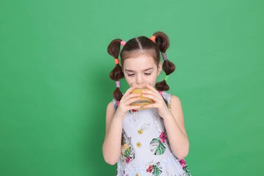 Sekiz yaşında, harika bir ruh hali içinde, mavi bluzlu, çiçekli, komik bir saç stili olan güzel bir kız yeşil arka planda şeffaf bir bardaktan portakal suyu içiyor.