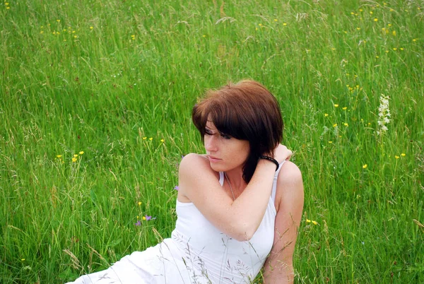 白いドレスを着た若い可愛い女の子が暑い夏の日に畑に座っている 彼女は左の手を肩に置き 髪をまっすぐにした 彼女は頭を横に向け遠くを見る — ストック写真