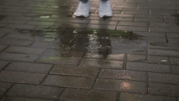 Mujer con jeans rotos y zapatillas blancas salta a un charco en la acera — Vídeo de stock