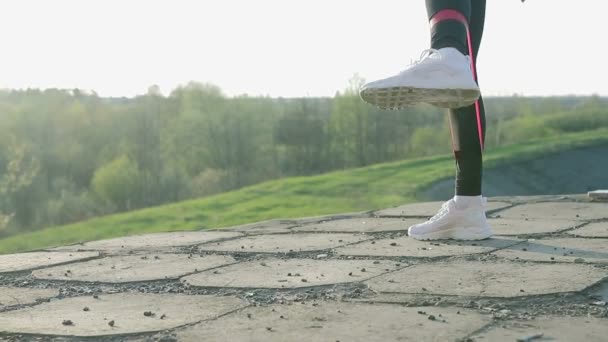 Joven linda chica haciendo gimnasia al atardecer. El atleta realiza ejercicios con una banda elástica de fitness — Vídeos de Stock