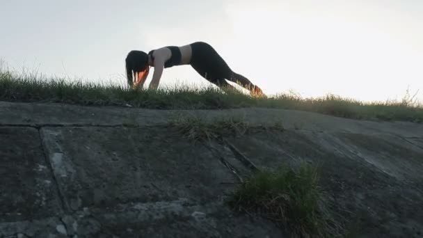 Joven bastante deportista se dedica a la aptitud en la naturaleza. La chica hace ejercicios en el énfasis que yace en el fondo de la puesta del sol — Vídeos de Stock