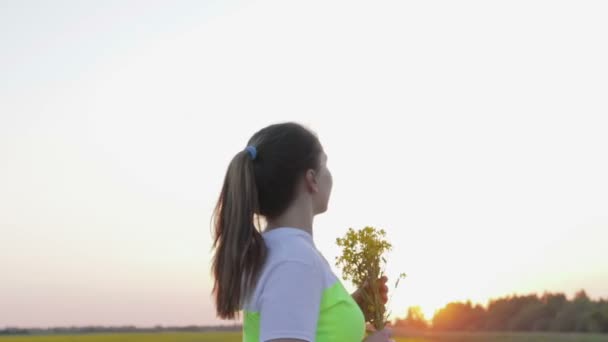 Uma jovem com um buquê de flores de canola caminha ao pôr do sol — Vídeo de Stock