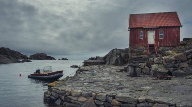 Landscape in Norway. solitude with nature. Traditional lonely Red wooden hut on the rocky shore of the northern sea and boat in the bay.