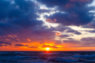 Gün batımında bulutlu bir gökyüzünde parlak turuncu güneş.