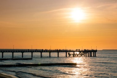 silhouettes of people walking on a pier by the sea against a sunset orange sky