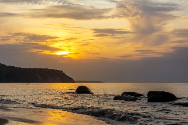 Arka planda bir uçurum, suda büyük kayalar, turuncu ışıkta gün batımında alçak güneş.