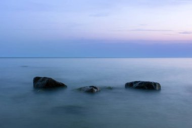 Alacakaranlık mavisi ışıkta sudan çıkan büyük kayalarla uzun ve pürüzsüz bir deniz yüzeyi.