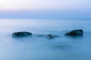 Alacakaranlık mavisi ışıkta sudan çıkan büyük kayalarla uzun ve pürüzsüz bir deniz yüzeyi.