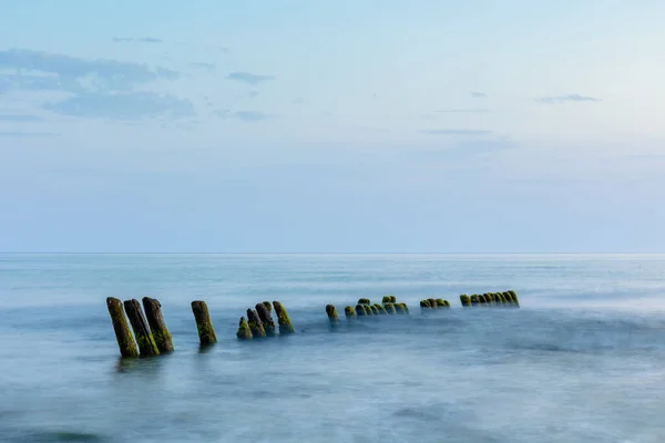 青い夕暮れ時に水から突き出た防波堤を持つ長い露出滑らかな海の表面 — ストック写真