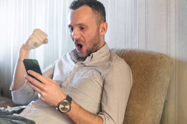 Kanepede oturan genç adam akıllı telefonun ekranına bakar ve sevinir.