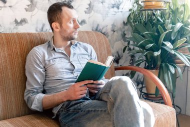 Genç adam kanepede oturmuş kitap okuyor ve ne yazık ki yan tarafa bakıyor. Koronovirüs kovid 19 karantinası sırasında evde yalnızlık çekiyor.