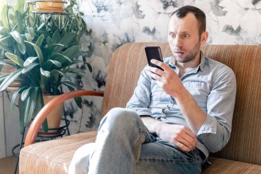 Korkan genç adam, salgın sırasında kanepede tek başına otururken akıllı telefondan haberleri okuyor.