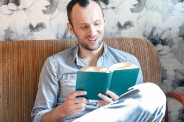 Gülümseyen genç bir adam bir dairede kanepede otururken kitap okur.