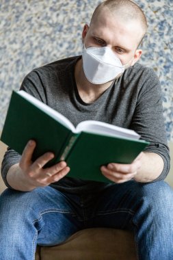 Coronavirus covid-19 karantinası sırasında evinde kendini izole eden ffp1 solunum cihazına sahip genç bir adam bir kitap okuyor.
