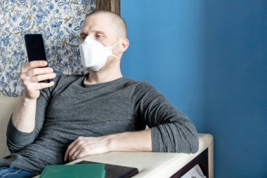 Coronavirus covid-19 karantinası sırasında evinde kendini izole eden ffp1 solunum cihazına sahip genç bir adam akıllı telefondan haberleri okuyor.