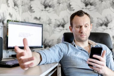 Genç bir erkek bilgisayar başında şaşkın bakışlı bir akıllı telefondan haberleri okuyor.