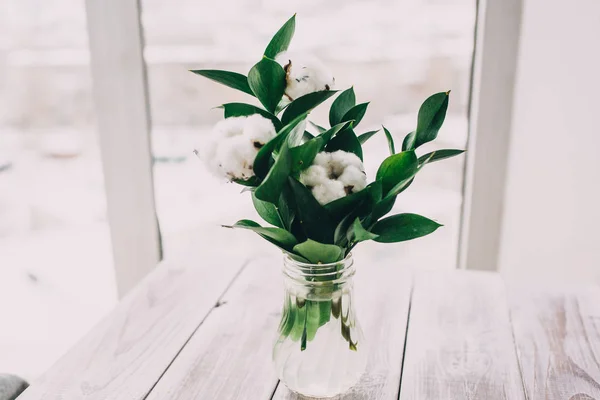 bouquet of cotton in a glass vase on light wooden background and the window