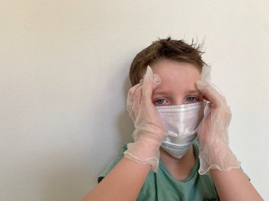 Evde üzgün bir çocuk var. Karantinada, eldiven giyiyor ve koruyucu bir maske takıyor.