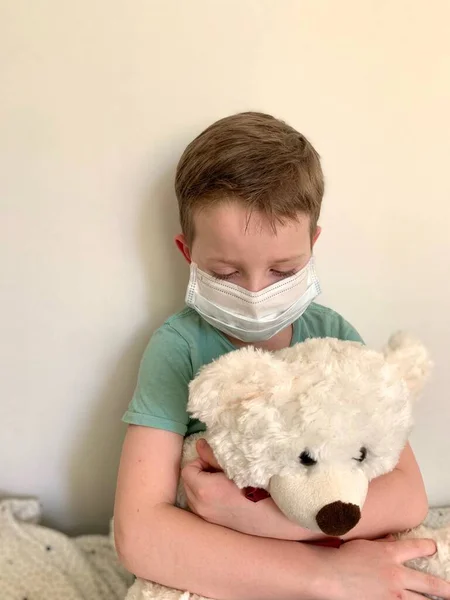 Een kleine, droevige jongen in een t-shirt, zittend met een smet in zijn handen, en een beschermend masker tegen virussen en ziektes. — Stockfoto