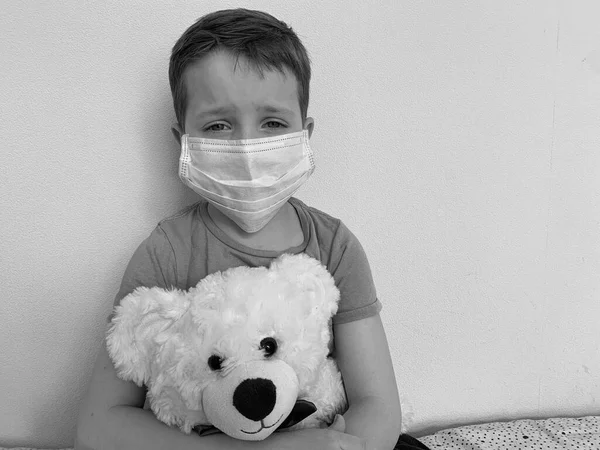 Een kleine, droevige jongen in een t-shirt, zittend met een smet in zijn handen, en een beschermend masker tegen virussen en ziektes. — Stockfoto