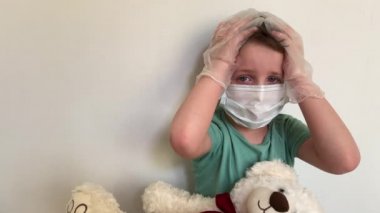 Maskeli, lateks eldivenli, oyuncak ayıyla ağlayan bir çocuk kameraya bakar..
