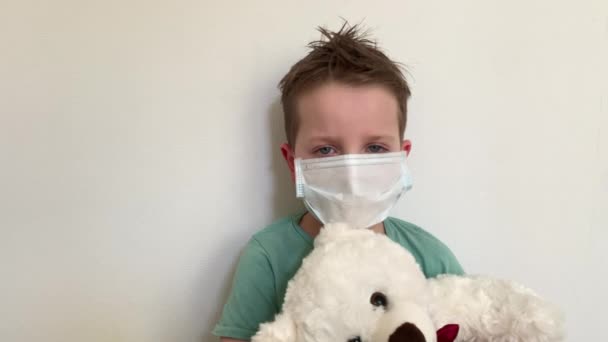 Un bambino con una maschera protettiva siede con un orsacchiotto, a casa in quarantena. guardando la telecamera, lampeggiando . — Video Stock