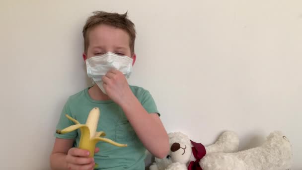Un niño pequeño con una máscara protectora de virus y enfermedades, comiendo un plátano, en el fondo de un osito de peluche . — Vídeo de stock
