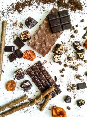 Değişik çikolata çeşitleri, beyaz bir masaya serpilmiş çikolata tüpleri kakao tozu, kahve çekirdekleri, kurutulmuş kayısılar.