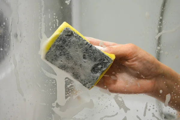 A womans hand with a yellow sponge washes the glass in the shower stall