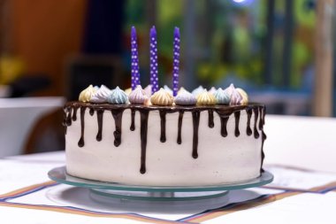 Doğum günü partisinde masada mor mumlar ve yemek süslemeleri olan doğum günü pastası: beyaz krema, çikolata ve mavi, pembe ve sarı kremalar evdeki masada.