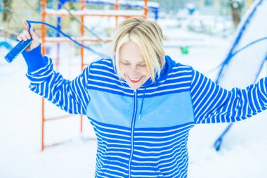 Aktif, sarışın bir kadın antrenman yapıyor ve ısınma egzersizini açık havada, karlı havada ip atlarken yapıyor.