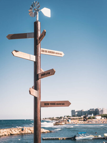 Wooden Route pointer showing way to cities of world with handmade wind-vane on top and beach on background.