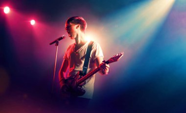 Genç sanatçı mikrofonla şarkı söyler ve sahnede gitar çalar. Müzik performansı