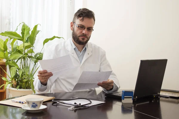 Médico en la oficina sentado en un escritorio y leyendo registros médicos — Foto de Stock