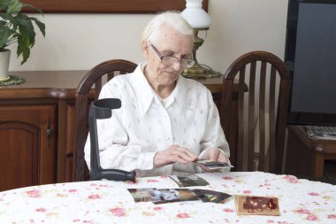 Old woman sitting in the living room at the table and looks at old photographs.  clipart