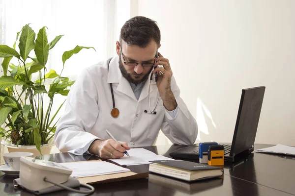 El médico se sienta en su escritorio, tomando notas hablando por teléfono . — Foto de Stock