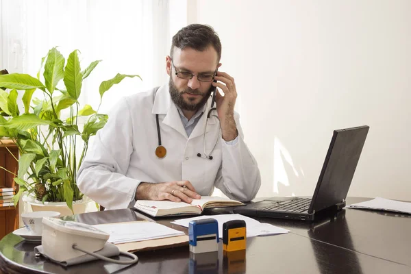 Médico en el consultorio del médico hablando por teléfono y comprobando las fechas disponibles en el calendario . — Foto de Stock