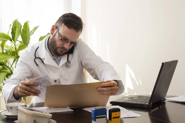 Un médico sentado en su escritorio. Hablando por teléfono revisando las historias clínicas del paciente — Foto de Stock