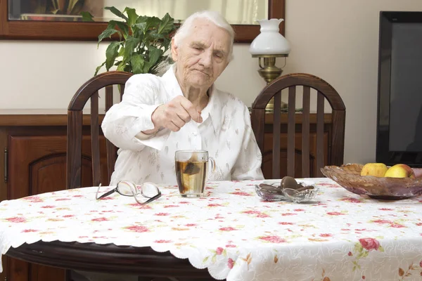 La anciana se sienta en una mesa en la sala de estar y hace té en un vaso. . — Foto de Stock