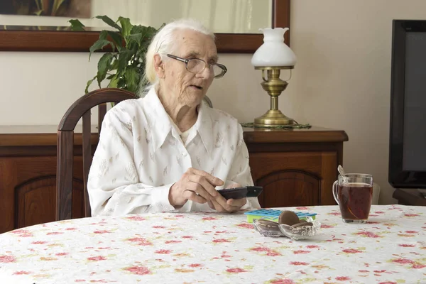 The old woman turns on the TV remote control sitting at the table. — Stock Photo, Image