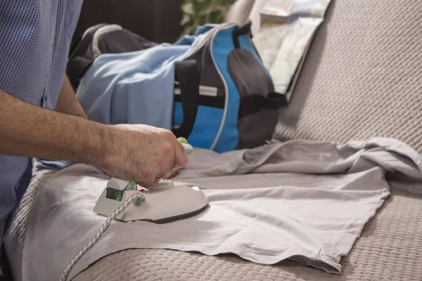 Ironing clothes on vacation. The man irons clothes with a tourist iron. The T-shirt and travel bag lie on the couch.