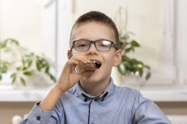 Gülümseyen okul çocuğu masada oturuyor. Elinde dişlerini ısıran bir çikolata tutuyor..