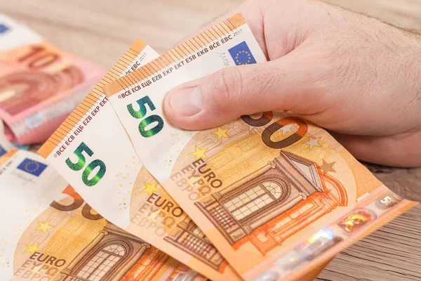 A man\'s hand holds two 50 Euro notes. In the background, euro banknotes lie on the table.