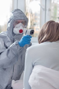 Koruyucu giysili, koruyucu maskeli ve gözlüklü biri lazer termometreyle ısıyı kontrol ediyor. Termometre tek kullanımlık eldiven takıyor..