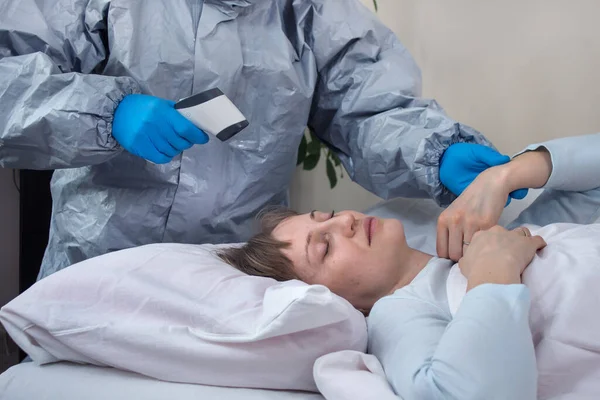 那个生病的女人躺在床上 穿着防护服和橡胶手套的人用激光温度计测量妇女的体温 另一方面 他测量病人的脉搏 帮助检疫人员 — 图库照片
