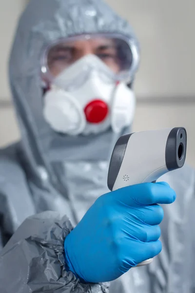 In the foreground a hand wearing a disposable glove holds a laser thermometer and measures the temperature. In the background, a figure wearing a protective suit, face mask and safety glasses.