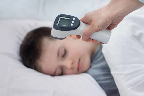 In the foreground a hand holds an electronic thermometer ready for measurement. In the background the face of a boy sleeping in bed on white bedding.