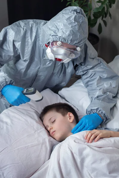 A sick school-age boy is lying in bed. A person wearing a protective suit and rubber gloves measures the child\'s temperature with a laser thermometer. With the other hand, he measures the pulse of a sick person. Help people in quarantine.